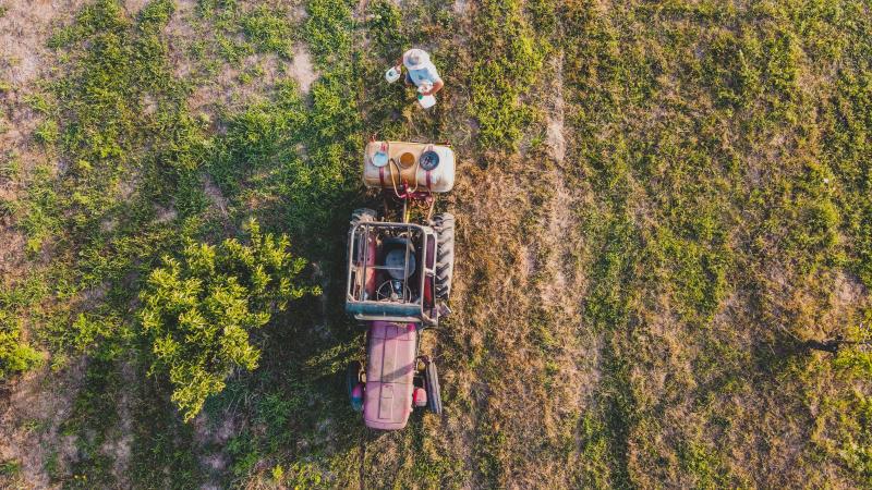 Image seen from above. A man with pesticide containers in his hand stands beside a tractor in a field.